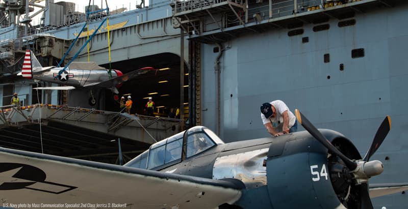 An FM-2 Wildcat being craned aboard an aircraft carrier while a crew member works on the aircraft in the foreground